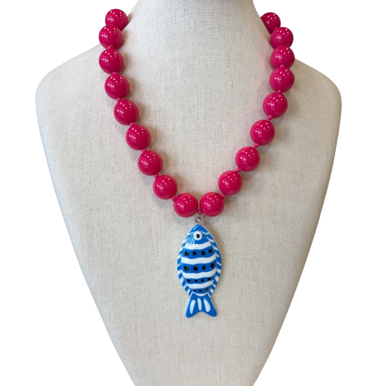 A bright pink beaded necklace displayed on a cream fabric jewelry bust. The necklace features glossy hot pink round beads with a central pendant shaped like a blue and white striped fish.