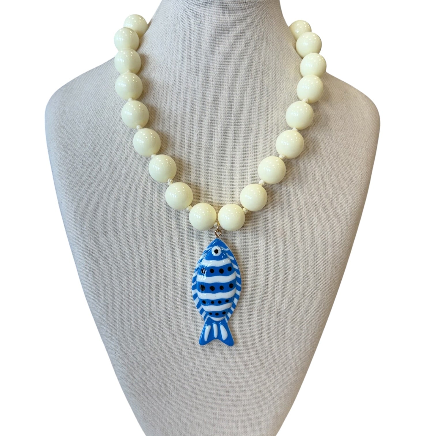A beaded necklace displayed on a fabric-covered jewelry bust. The necklace consists of round cream-colored beads with a blue and white striped fish pendant hanging from a gold-tone ring.