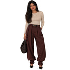 A woman modeling high-waisted, wide-leg brown pants with a matching belt, paired with a beige long-sleeve top, black heels, and a black woven handbag, set against a plain white background.