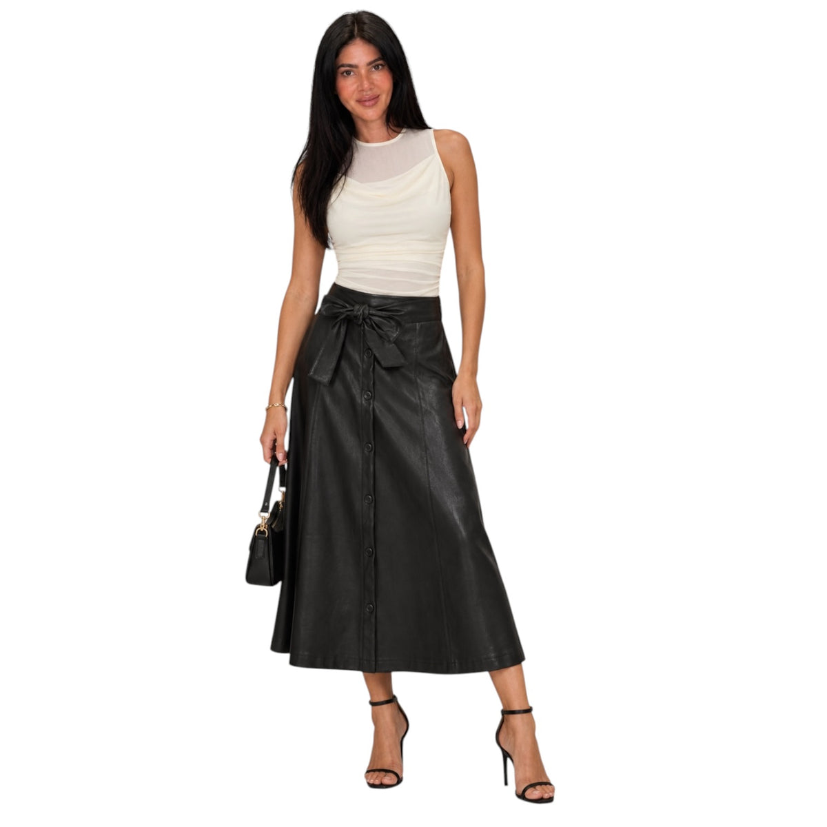 A woman modeling a black faux-leather midi skirt with a tie waist and button front, paired with a cream ribbed tank top and black high-heeled sandals, against a plain white background.