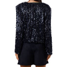 A model shown from the back wearing a long-sleeve, dark navy blue textured jacket covered in dense, shimmery silver and white fringe, paired with dark shorts against a plain white background.