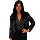 A woman modeling a black long-sleeve textured jacket with silver glitter-like flecks over an olive green cowl-neck top and black skirt, against a plain white background.
