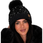 A close-up of a woman wearing a black knit beanie with a large black faux-fur pom-pom, black sequins, and a small brown UGG logo patch on the side.