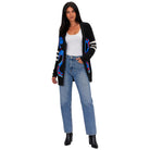 A woman modeling a black knit cardigan with colorful blue, red, and white graphic patterns on the front and sleeves, styled with a white tank top, blue jeans, and black ankle boots against a plain white background.