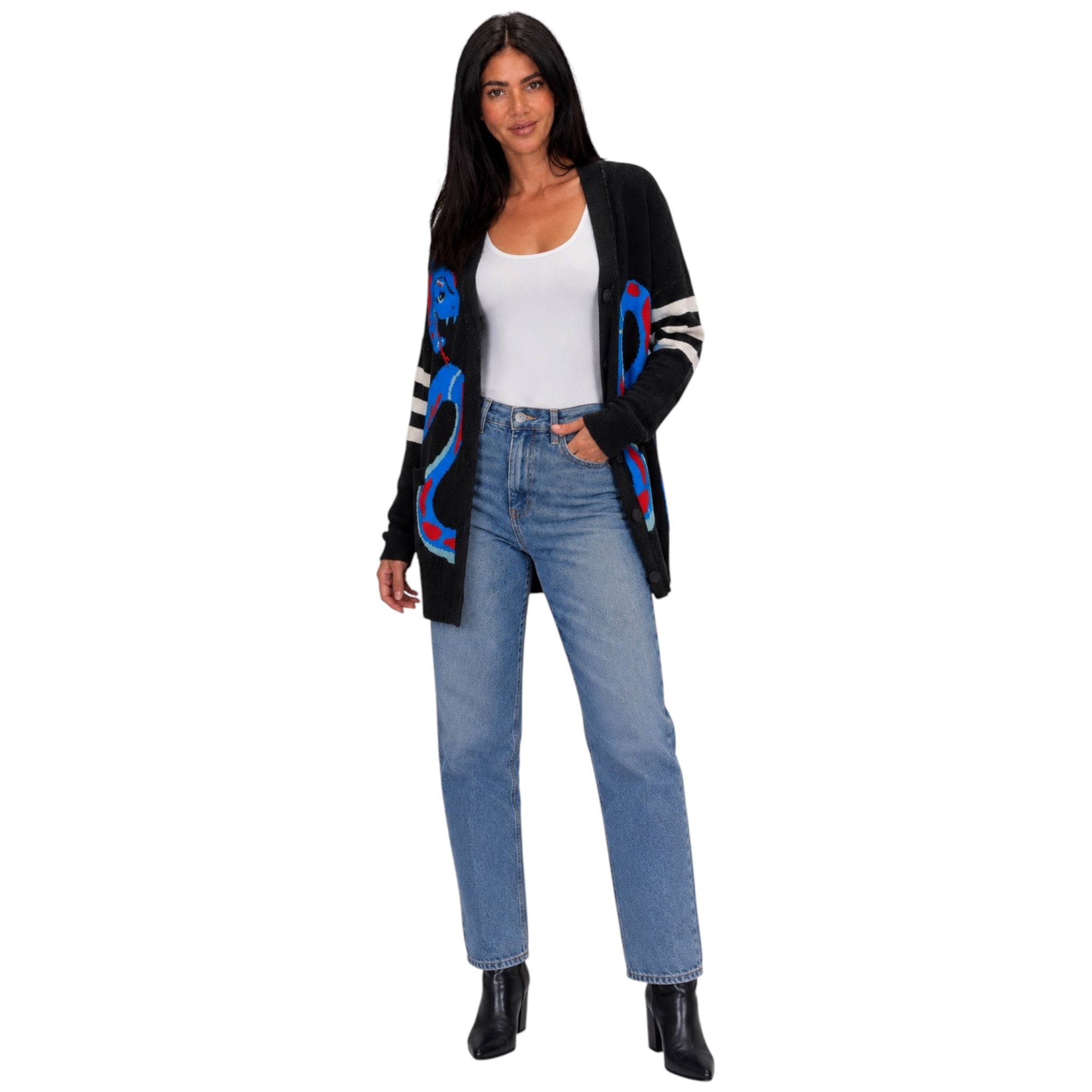 A woman modeling a black knit cardigan with colorful blue, red, and white graphic patterns on the front and sleeves, styled with a white tank top, blue jeans, and black ankle boots against a plain white background.