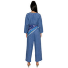 A woman viewed from the back wearing a blue denim jumpsuit with a patterned blue and white belt, set against a plain white background.