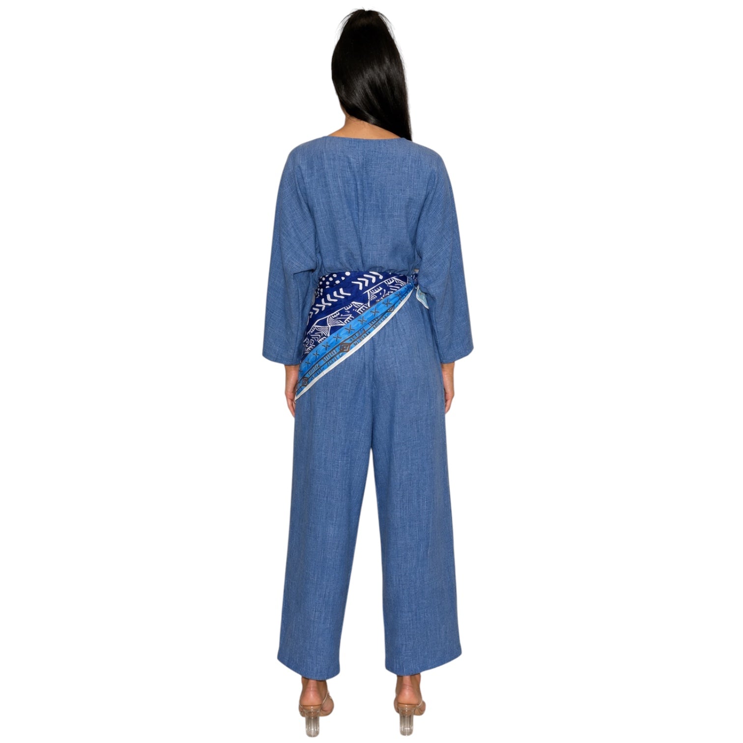A woman viewed from the back wearing a blue denim jumpsuit with a patterned blue and white belt, set against a plain white background.