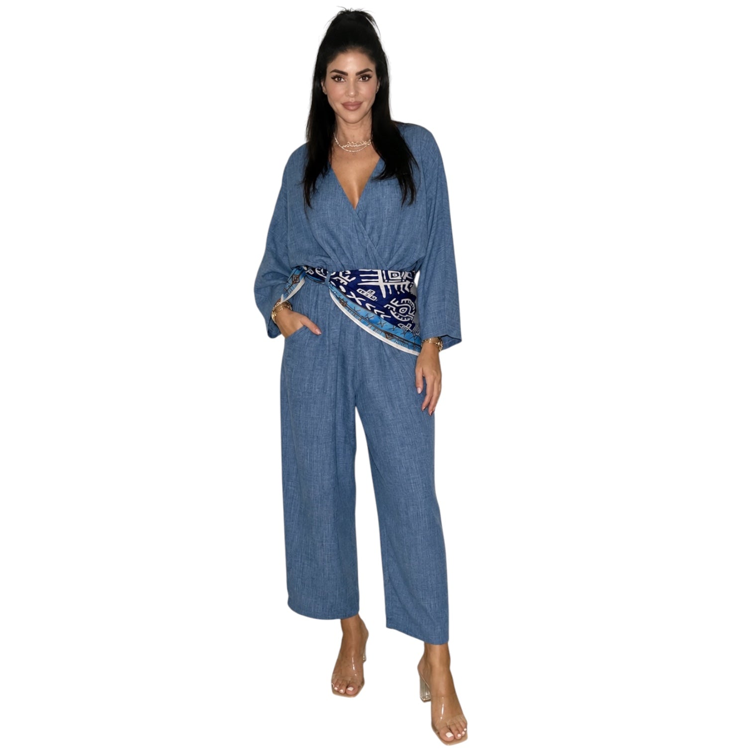 A woman modeling a blue denim-like jumpsuit with a patterned blue, white, and black waist tie, shown against a plain white background.