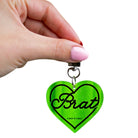 Green heart-shaped keychain with 'Brat' text held by a hand on a white background