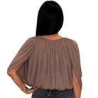 A woman with long black hair wearing a taupe-brown draped short-sleeve blouse and dark jeans, standing against a plain white background.