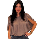 A woman with long black hair wearing a taupe-brown draped short-sleeve blouse and dark jeans, standing against a plain white background.
