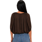 A woman viewed from the back wearing a dark brown, short-sleeved blouse with voluminous gathered sleeves and a wide neckline, paired with blue jeans against a plain white background.