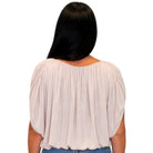 A woman viewed from the back wearing a light blush pink pleated blouse with short puff sleeves and a wide neckline, paired with blue jeans against a plain white background.