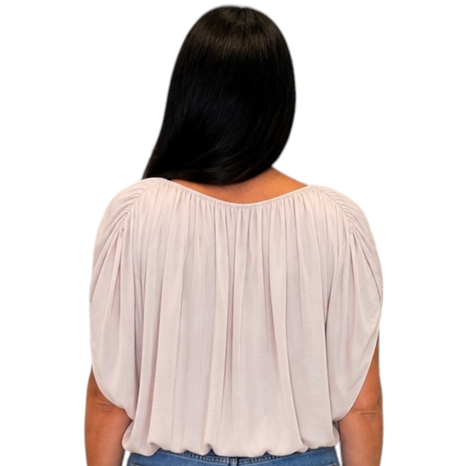A woman viewed from the back wearing a light blush pink pleated blouse with short puff sleeves and a wide neckline, paired with blue jeans against a plain white background.