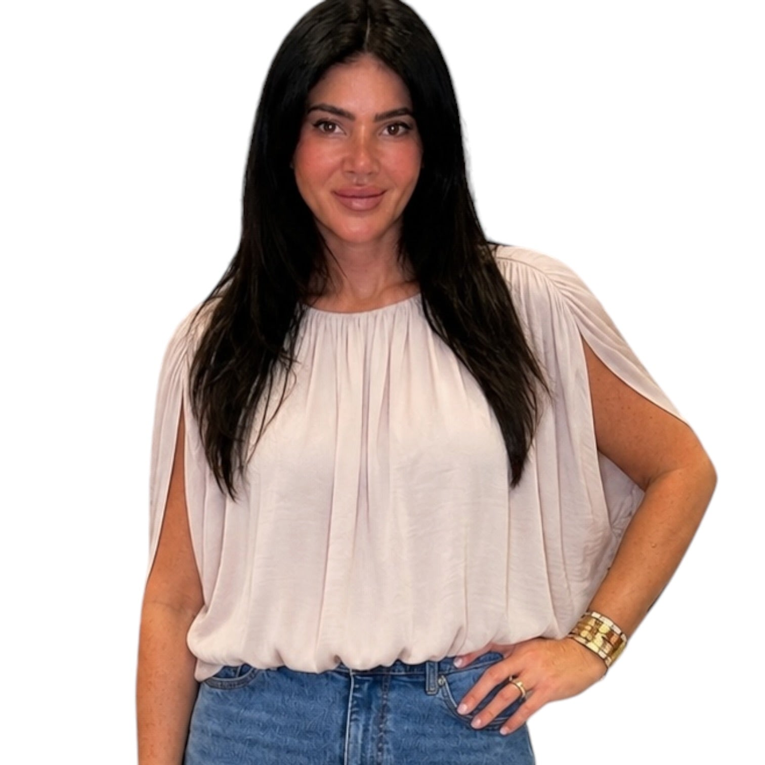 A woman with long dark hair wearing a light beige, short-sleeved top with draped sleeves and blue jeans, standing against a plain white background.