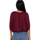 A woman viewed from the back wearing a burgundy pleated blouse with three-quarter puff sleeves and blue jeans, set against a plain white background.