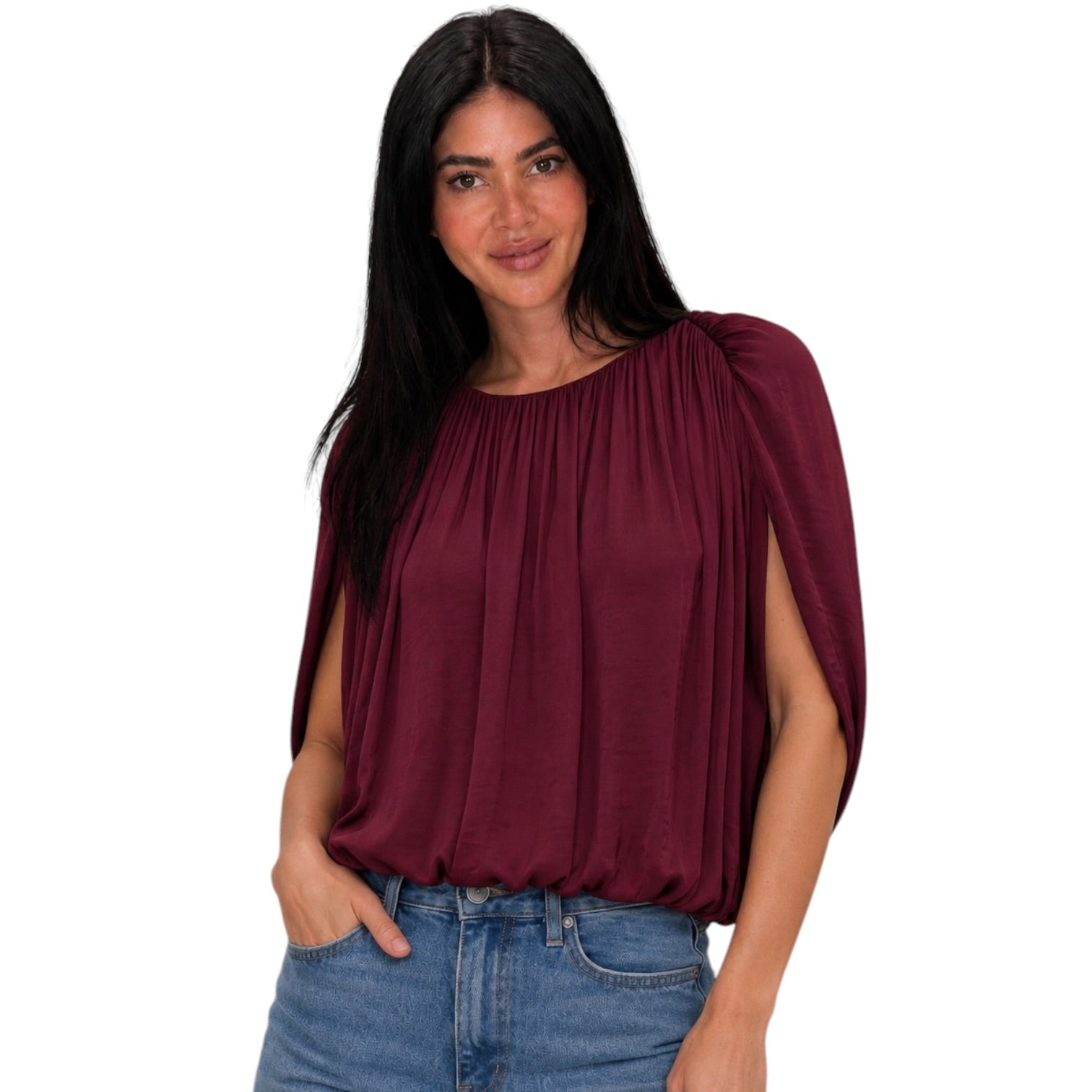 A woman wearing a draped, pleated top in deep burgundy with wide, cape-like sleeves, paired with blue jeans, against a plain white background.