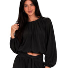 A woman wearing a black long-sleeve blouse with a gathered, balloon-style neckline and voluminous sleeves, paired with a matching black skirt, against a plain white background.