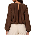 A woman shown from the back wearing a brown long-sleeve blouse with a keyhole back cutout and cream-colored pants, set against a plain white background.
