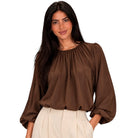 A woman wearing a brown long-sleeve blouse with a gathered, pleated neckline and cream-colored trousers, shown against a plain white background.