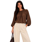 A woman modeling a brown long-sleeve blouse with a gathered neckline and pleated details, paired with cream wide-leg trousers, against a plain white background.