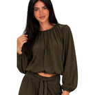 A woman wearing an olive green long-sleeve crop top with a gathered neckline and matching high-waisted tie-waist bottoms, shown against a plain white background.