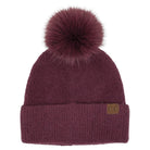 A dark burgundy knit beanie with a large matching faux fur pom-pom on top and a small brown leather 'CC' logo patch on the front cuff, shown against a plain white background.