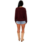 A woman shown from the back wearing a dark burgundy long-sleeve knit sweater, light blue denim shorts, and beige cowboy boots, set against a plain white background.