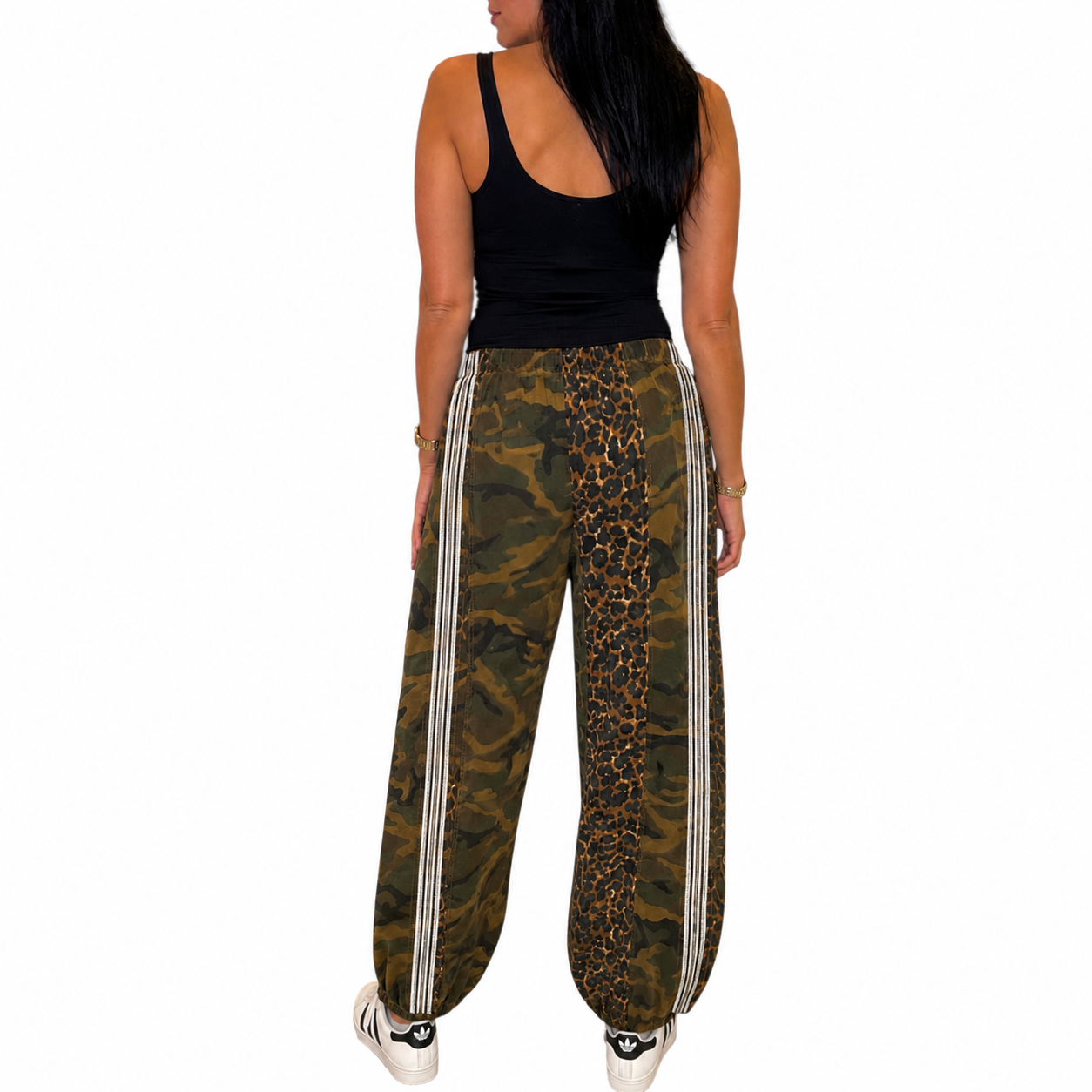 A woman shown from the back wearing wide-leg cargo-style pants with a mixed print of camouflage, leopard print, and vertical white stripes, paired with a black tank top and white sneakers on a plain white background.