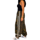 A woman modeling wide-leg harem pants with a green and brown camouflage pattern and leopard print side panels, paired with a black tank top and white sneakers, against a plain white background.