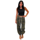 A woman modeling a pair of camouflage jogger pants with a white drawstring, paired with a black crop top, against a plain white background.