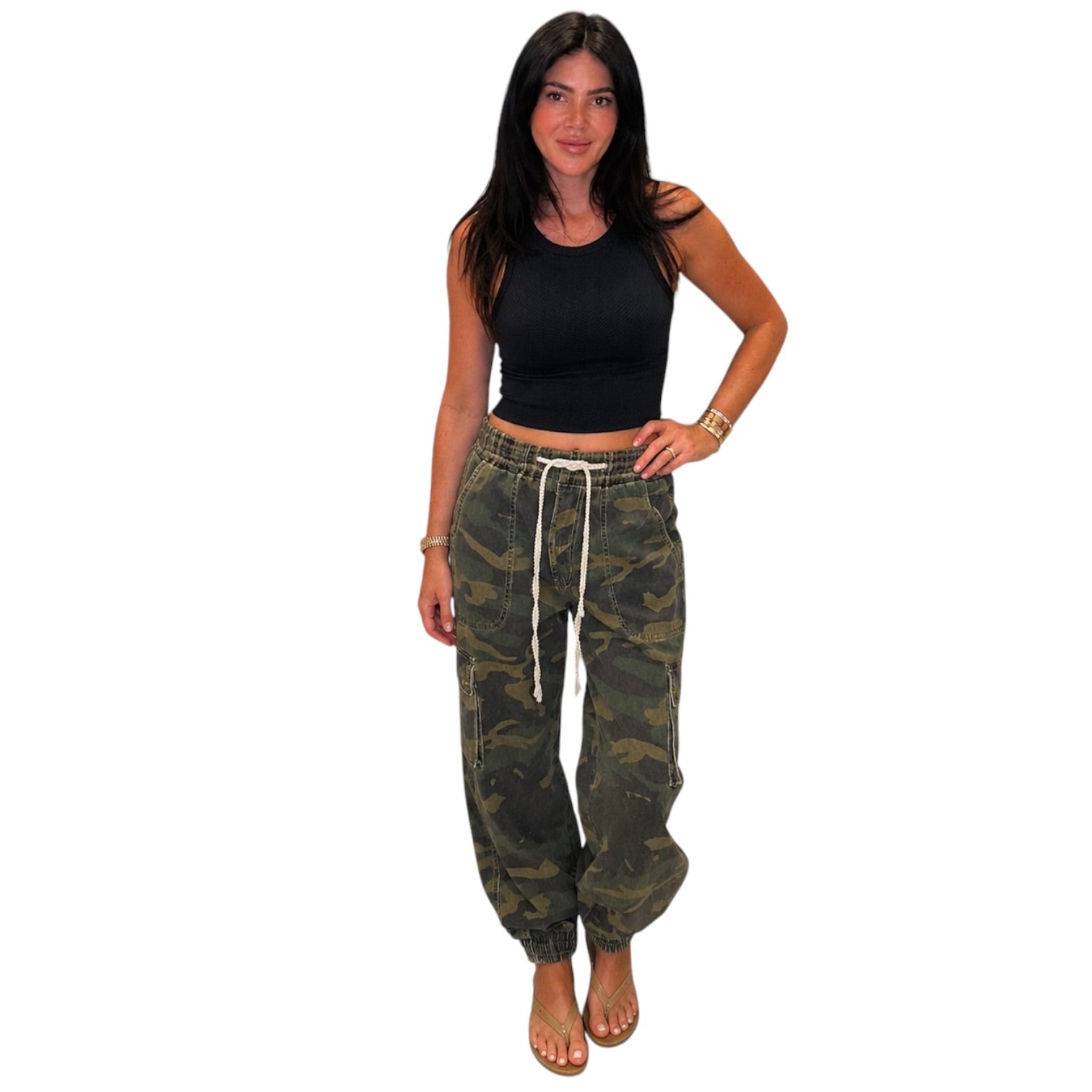 A woman modeling a pair of camouflage jogger pants with a white drawstring, paired with a black crop top, against a plain white background.