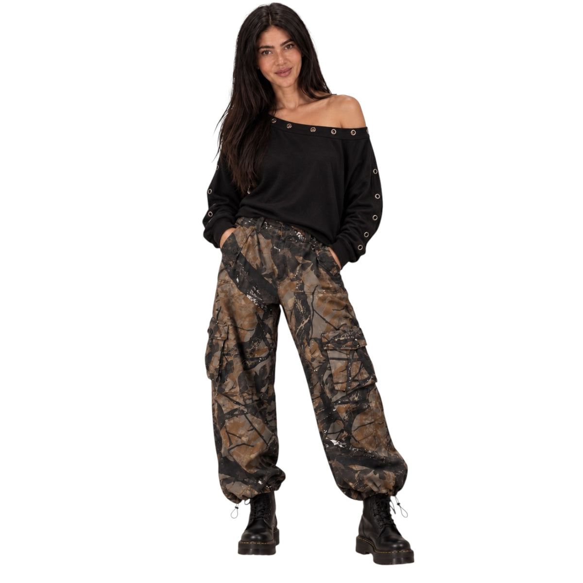 A woman modeling a black off-the-shoulder long-sleeve top with metal eyelets and brown camouflage cargo pants, paired with black combat boots, against a plain white background.