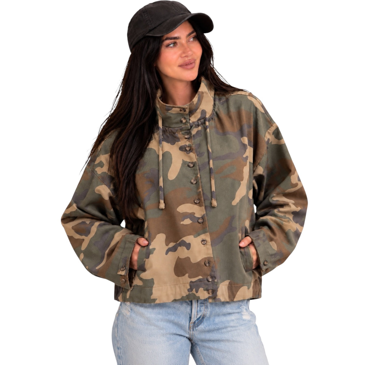 A woman wearing a camouflage jacket with a high collar and button front, paired with light blue jeans and a black cap, against a plain white background.