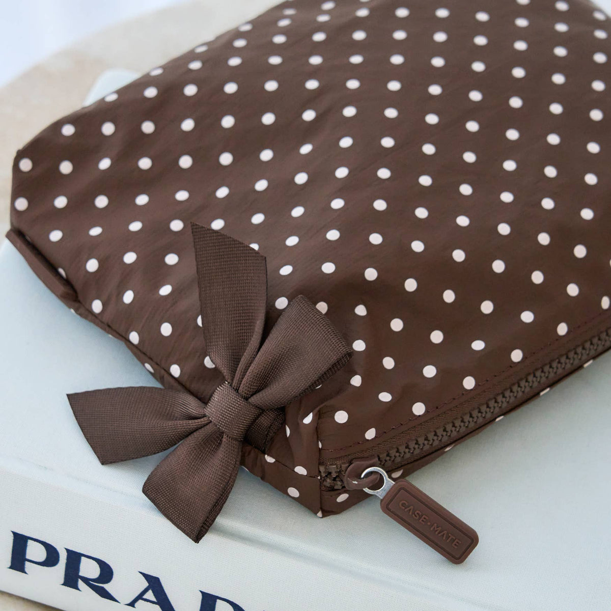 A brown nylon pouch with white polka dots, featuring a matching brown ribbon bow and a zip closure with a small brown pull tab, resting on a white surface.