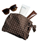 A brown cosmetic pouch with white polka dots and a matching bow, styled on a wooden surface with sunglasses, a tube of cream, and a brown tube.