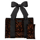 A structured tote bag with a glossy tortoiseshell pattern in brown and black, featuring a large matching black satin bow on the handle and a small matching zip pouch placed in front, shown on a plain white background.