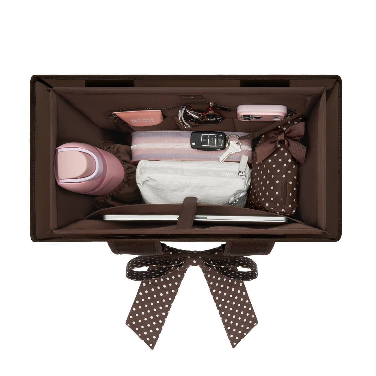 A brown organizer box shown from above, packed with travel accessories including a pink water bottle, a striped pouch, a white pouch, sunglasses, a pink case, and a polka-dot bag, on a white background.