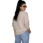 A woman with long black hair in a ponytail, wearing tortoiseshell sunglasses, a light beige knit sweater, and light blue jeans, shown from a back-three-quarter view against a plain white background.