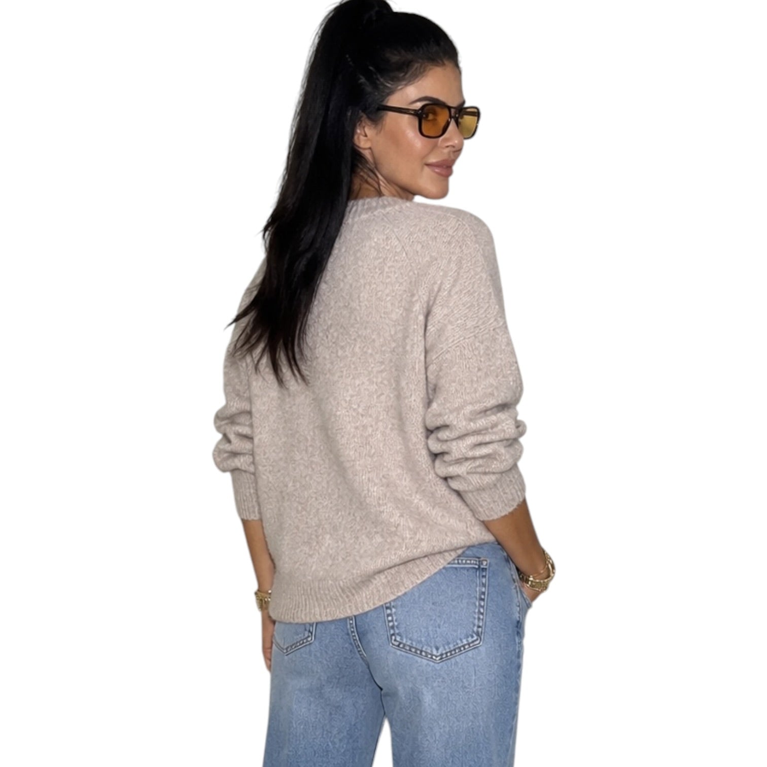 A woman with long black hair in a ponytail, wearing tortoiseshell sunglasses, a light beige knit sweater, and light blue jeans, shown from a back-three-quarter view against a plain white background.