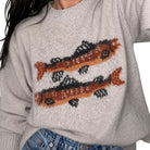 A close-up of a person wearing a light heather gray knit sweater with two large fish motifs in orange, black, and white across the chest, paired with blue jeans.