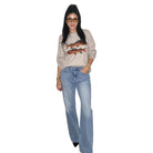 A woman modeling a beige crewneck sweatshirt with a fish graphic and light-wash blue jeans, standing against a plain white background.