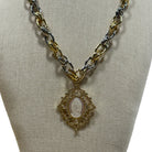 A two-tone gold and silver chain necklace with a gold pendant featuring a white oval religious medallion, displayed on a beige fabric jewelry bust.