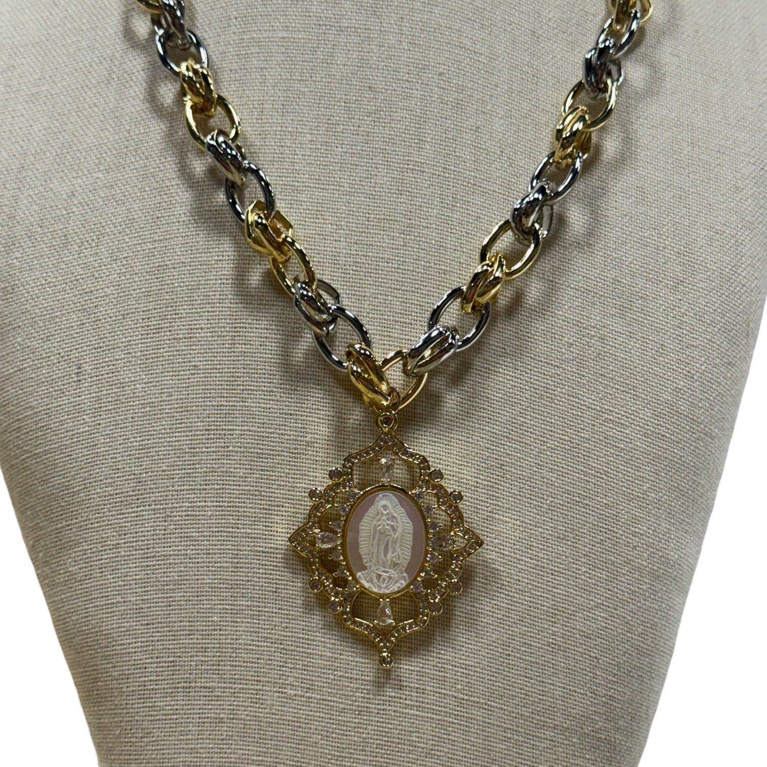 A two-tone gold and silver chain necklace with a gold pendant featuring a white oval religious medallion, displayed on a beige fabric jewelry bust.