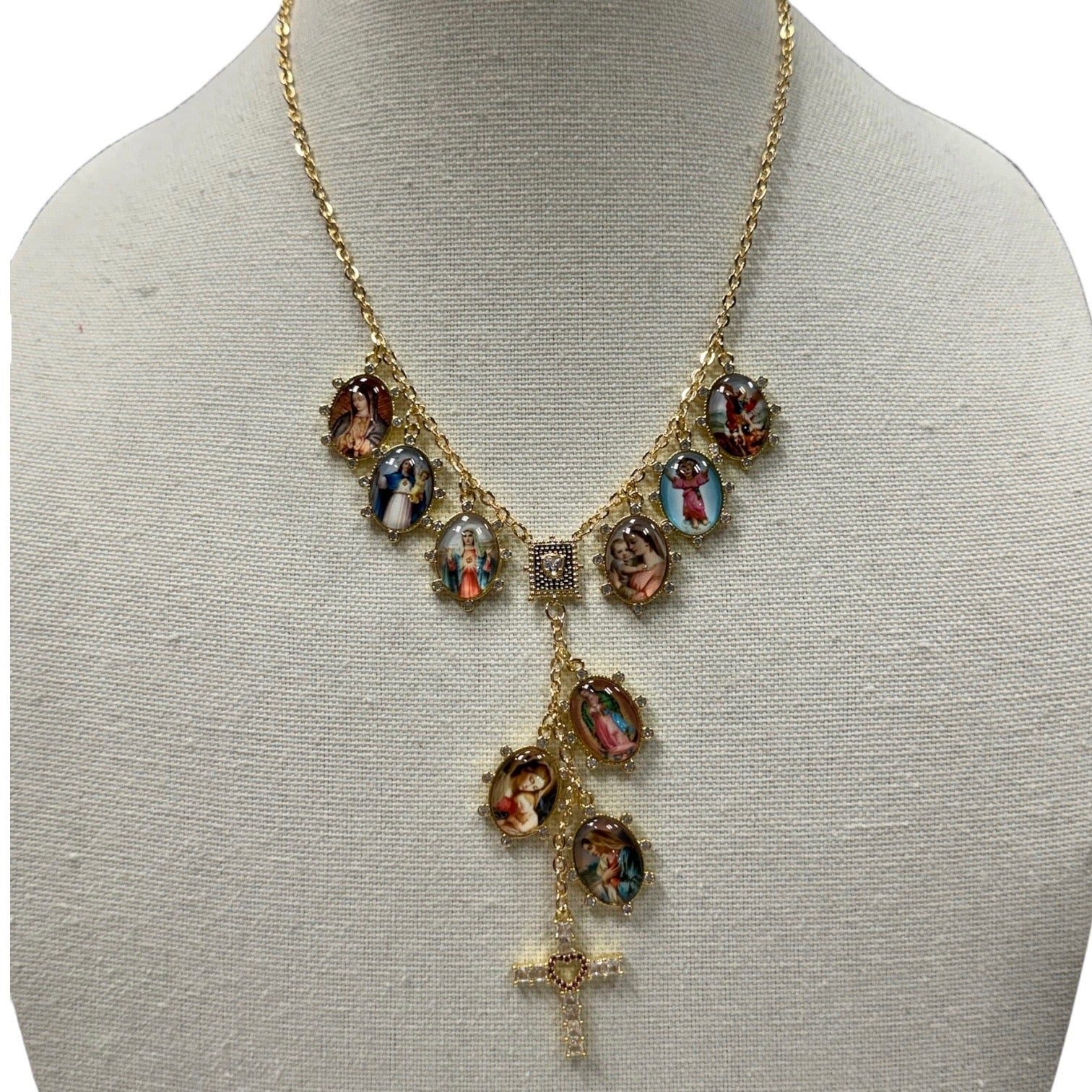 A gold-tone necklace displayed on a cream fabric mannequin bust. The necklace features a chain with multiple oval religious medallions depicting the Holy Family (Jesus, Mary, and Joseph) and a dangling cross at the bottom.