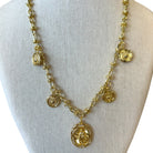 A gold-toned statement necklace displayed on a cream fabric jewelry bust. The necklace features a textured chain with multiple dangling charms, including oval medallions, floral links, and circular discs, with a large oval pendant at the center.