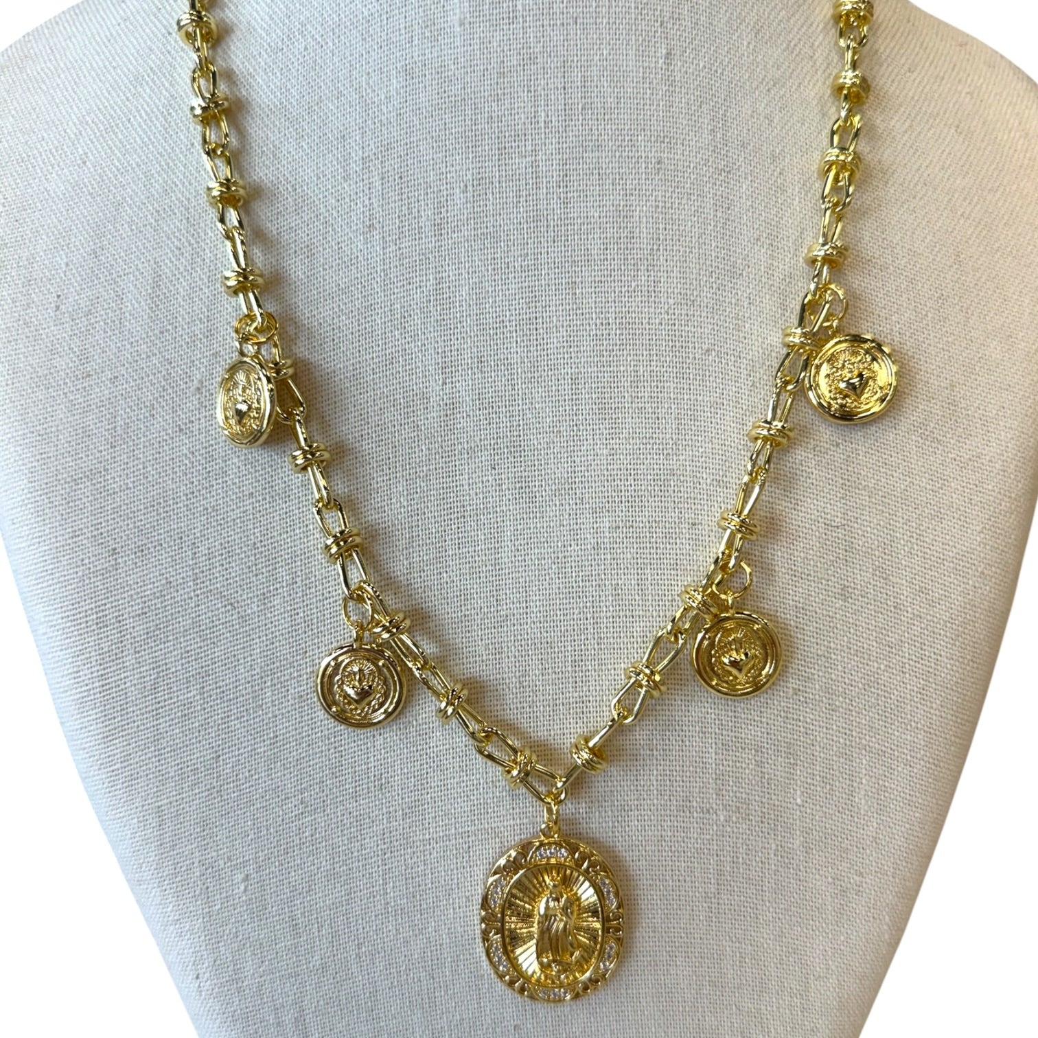 A gold-toned statement necklace displayed on a cream fabric jewelry bust. The necklace features a textured chain with multiple dangling charms, including oval medallions, floral links, and circular discs, with a large oval pendant at the center.