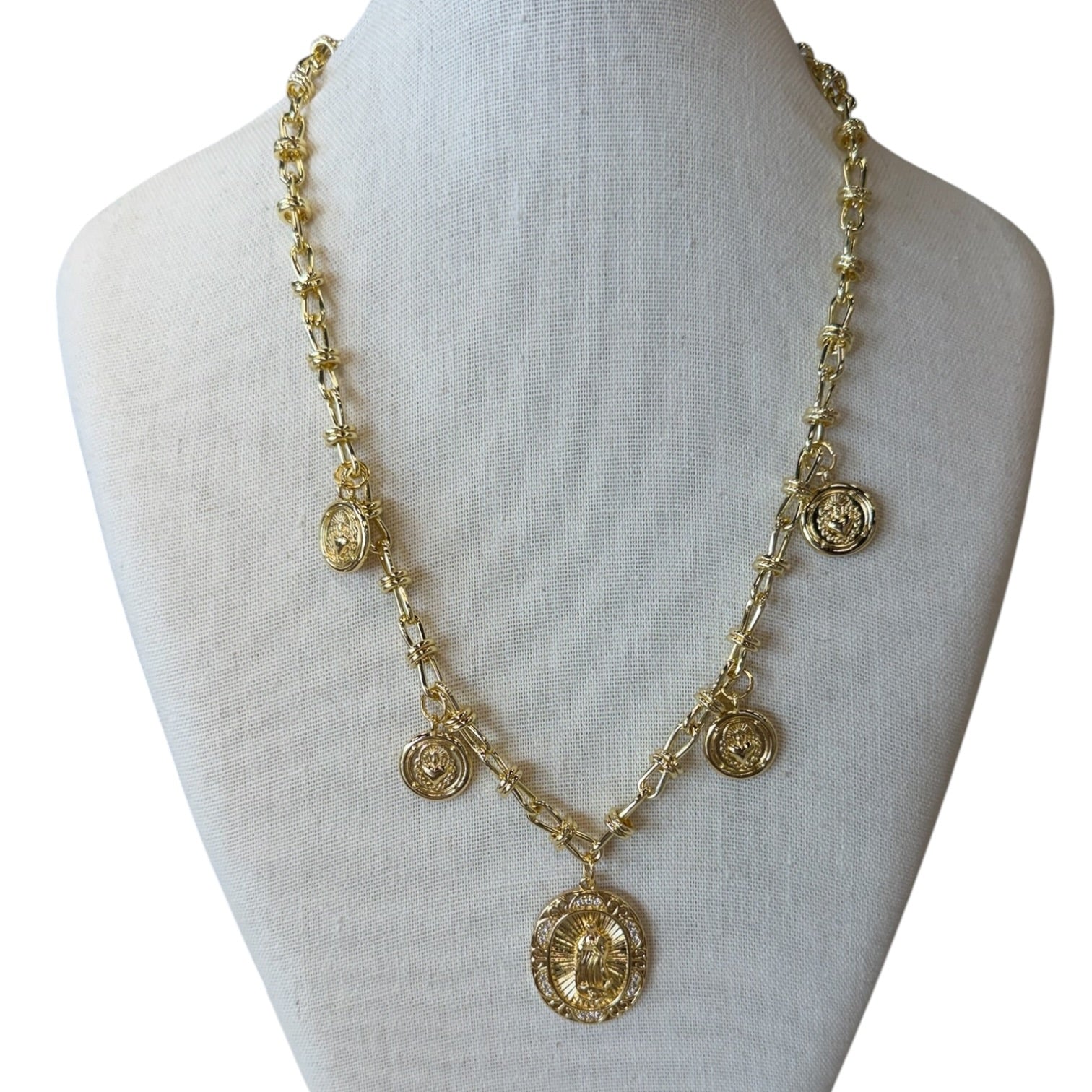 A gold-toned chain necklace displayed on a light beige fabric mannequin bust. The necklace features multiple small oval medallion charms and one larger central pendant with embossed detailing.