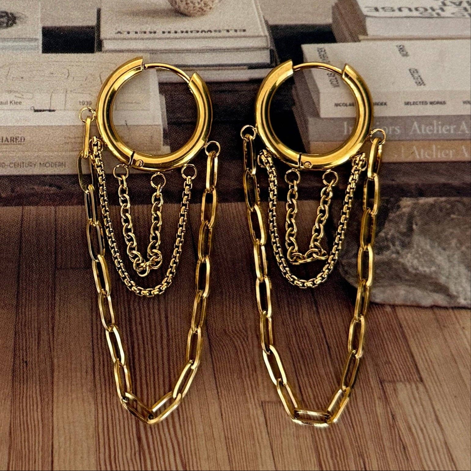 Gold hoop earrings with chain details on a wooden surface with books in the background