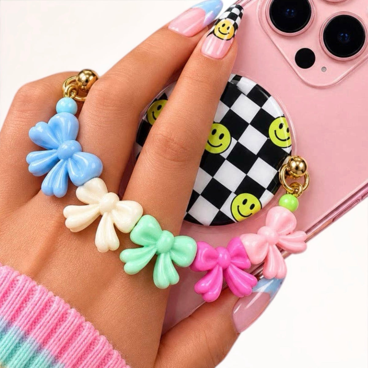 A close-up of a hand holding a pink smartphone, showing a decorative wrist strap with five colorful bow charms in blue, cream, mint, pink, and light pink, plus gold-tone rings and beads. The phone case has a black-and-white checkered pattern with yellow smiley faces.